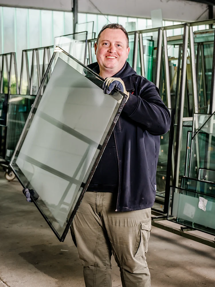 Glasermeister Marcel Plum als sympathischer Inhaber von Glas Plum aus Jülich steht in seinem Lager, hält Spezialglas in der Hand und lächelt in die Kamera.